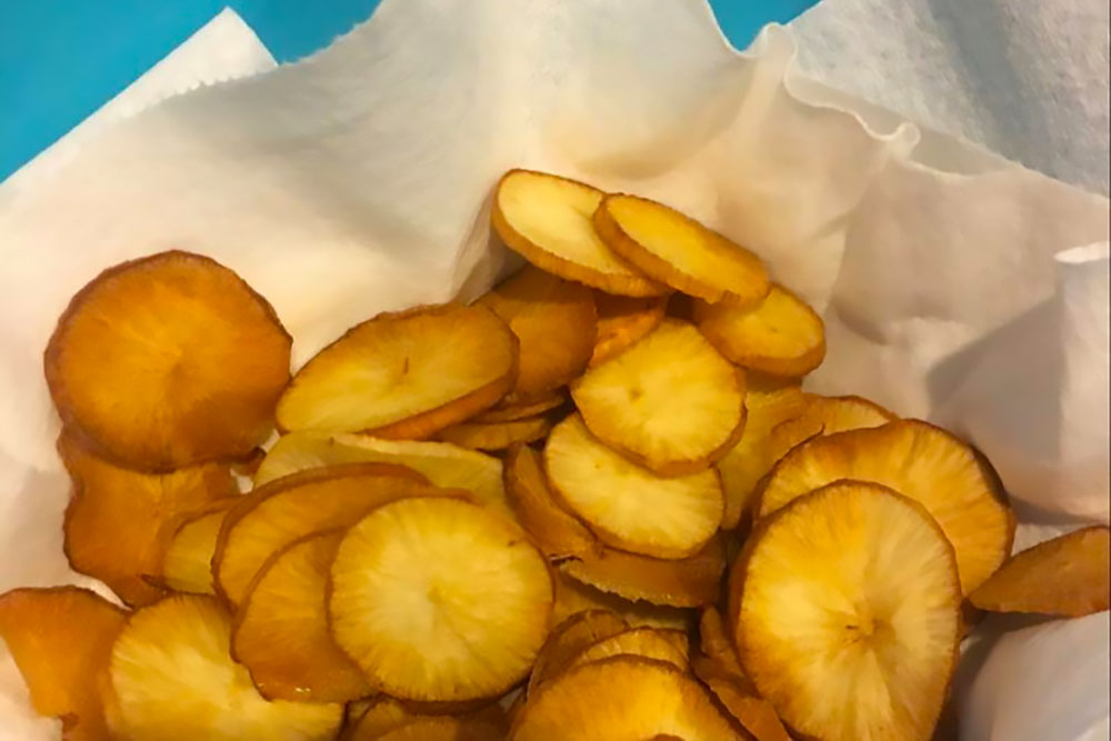 A basket of golden-brown, crispy plantain chips is set against a blue background, showcasing a vibrant, allergen-friendly snack option. These natural chips are perfect for those seeking fresh flavors and healthy living.| Daré Vita Café | Las Vegas