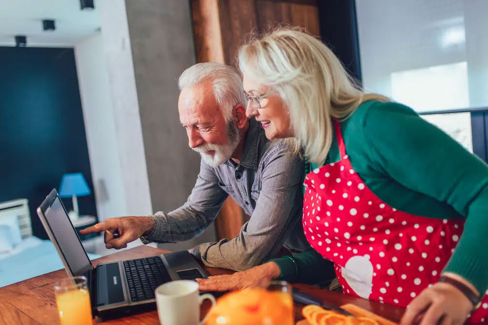 How-Can-Online-Training-for-Healthy-Eating-Help An elderly couple, with the woman in a red polka-dot apron, smiles while looking at a laptop in a cozy kitchen, featuring fresh orange slices, suggesting a wholesome and healthy living environment.| Daré Vita Café | Las Vegas