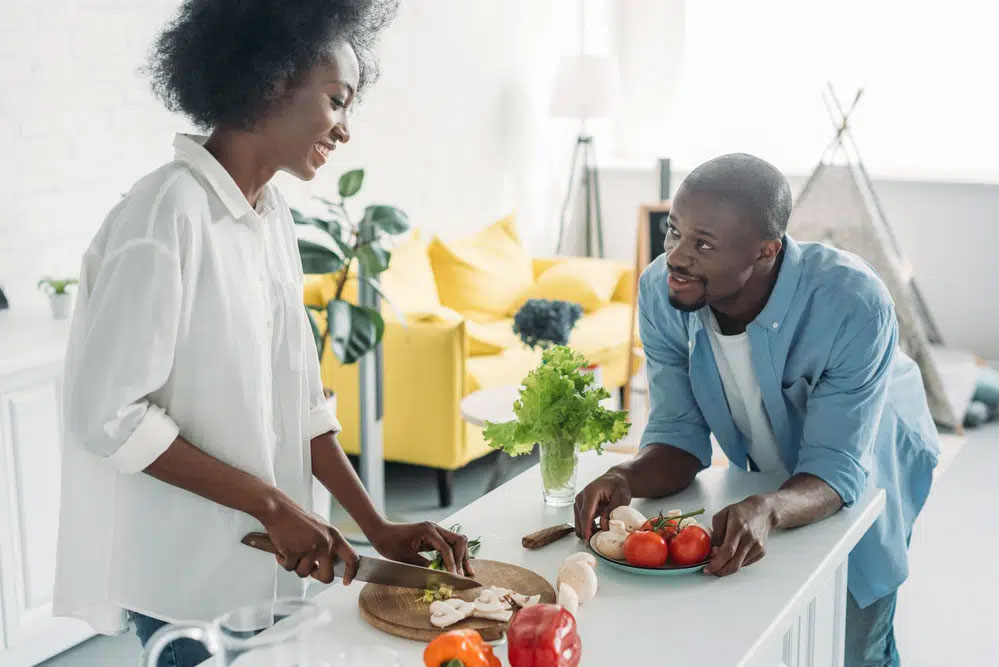 A woman in a white shirt chops mushrooms while a man in a blue shirt leans on the counter with tomatoes and fresh greens, creating a vibrant, wholesome meal. The kitchen's bright, modern style complements their healthy living focus.| Daré Vita Café | Las Vegas