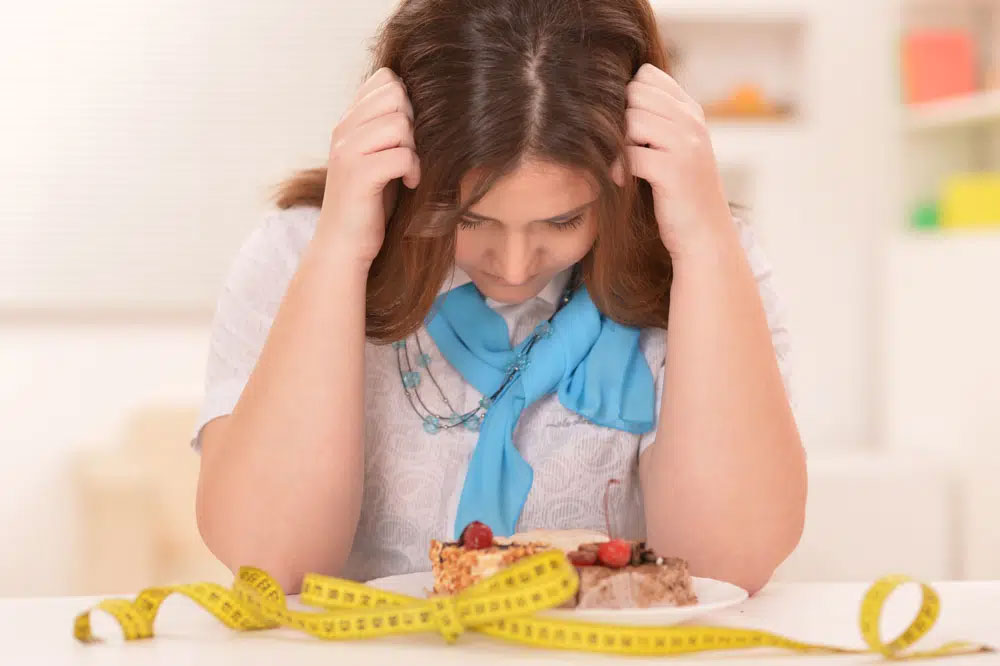 Why-diets-arent-healthy A woman with a blue scarf looks down at a cake on a plate, surrounded by a measuring tape, symbolizing food sensitivity. The scene highlights the importance of allergen-friendly and healthy living choices.| Daré Vita Café | Las Vegas