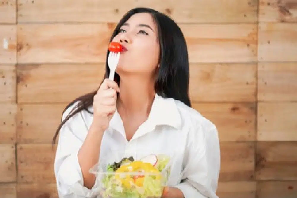 Get-Americans-Eating-Healthier A woman in a white shirt enjoys a fresh, vibrant salad with a cherry tomato on a fork. She is smiling, suggesting the meal is…| Daré Vita Café | Las Vegas