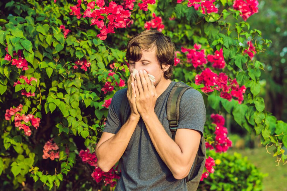 A person with brown hair and a backpack sneezes into a tissue in front of vibrant pink flowering bushes, highlighting the importance of allergen-friendly and natural environments for healthy living and food sensitivity awareness.| Daré Vita Café | Las Vegas