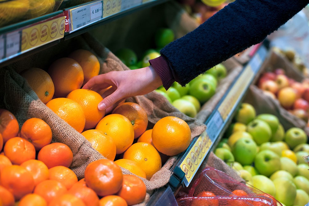 A person selects fresh oranges from a grocery store display filled with vibrant citrus and apples, emphasizing fresh produce and healthy living.| Daré Vita Café | Las Vegas