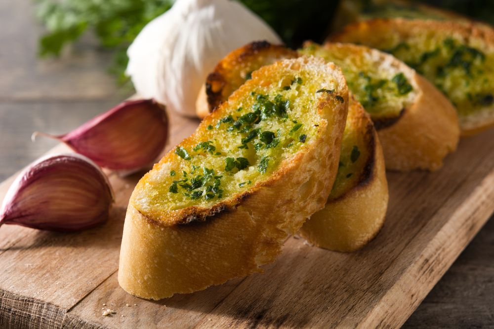 Slices of golden-brown garlic bread with vibrant green parsley sit on a wooden board, accompanied by fresh garlic cloves, embodying fresh flavors and wholesome, natural ingredients for nourishing meals.| Daré Vita Café | Las Vegas