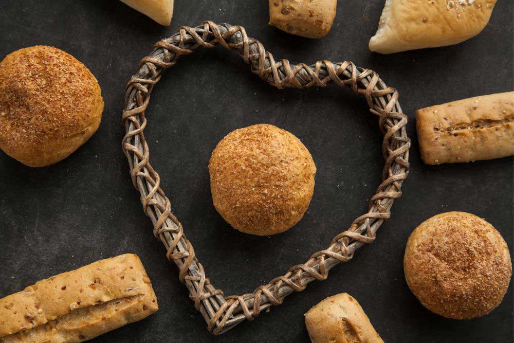 A heart-shaped wicker frame surrounds a round, golden-brown bread roll on a dark surface, surrounded by various other breads. The image highlights the natural, wholesome ingredients used for allergen-friendly baking.| Daré Vita Café | Las Vegas