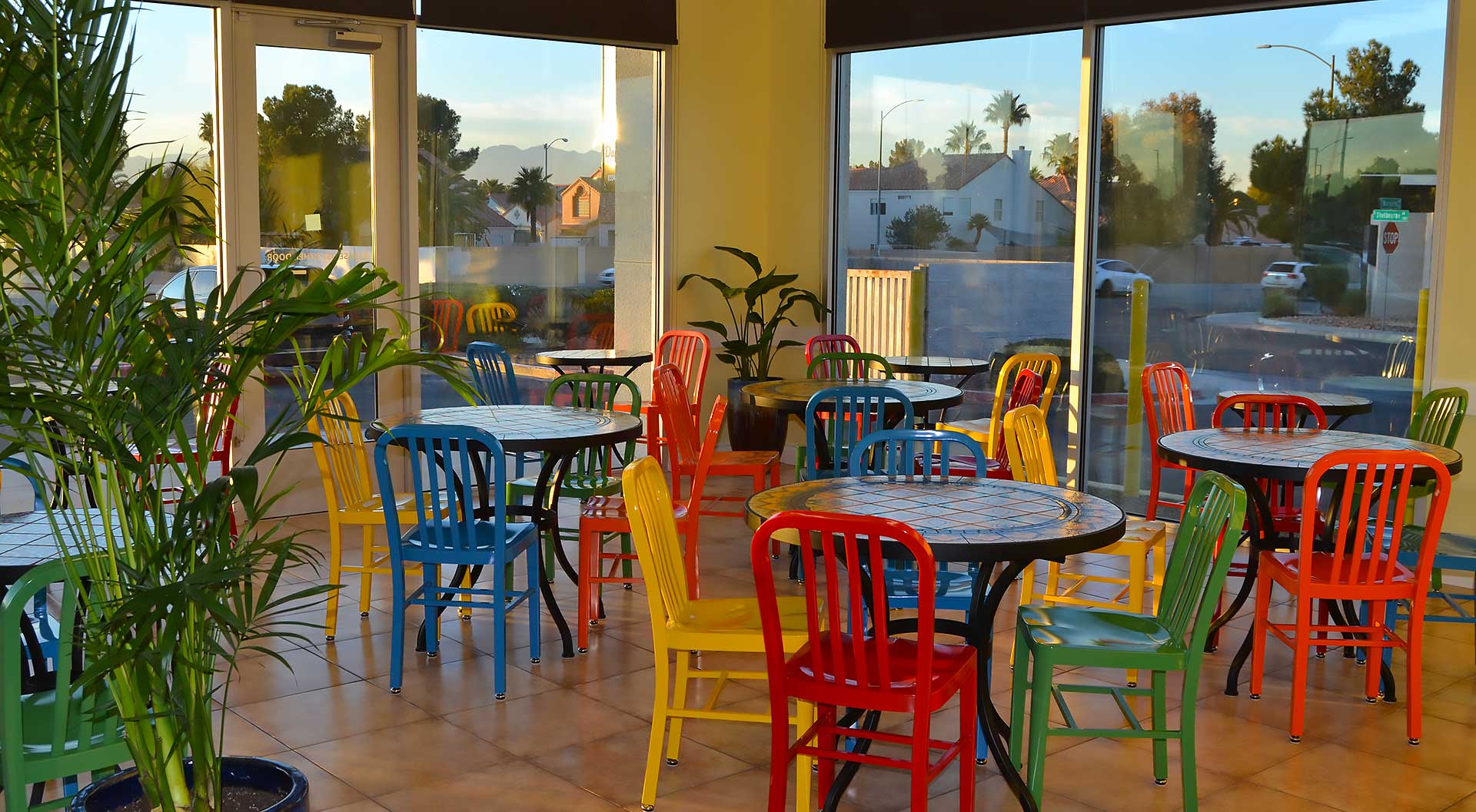 A vibrant café interior with colorful chairs and tables, surrounded by large windows letting in natural light. Green plants add to the fresh ambiance, reflecting a commitment to allergen-friendly, wholesome meals and healthy living.| Daré Vita Café | Las Vegas