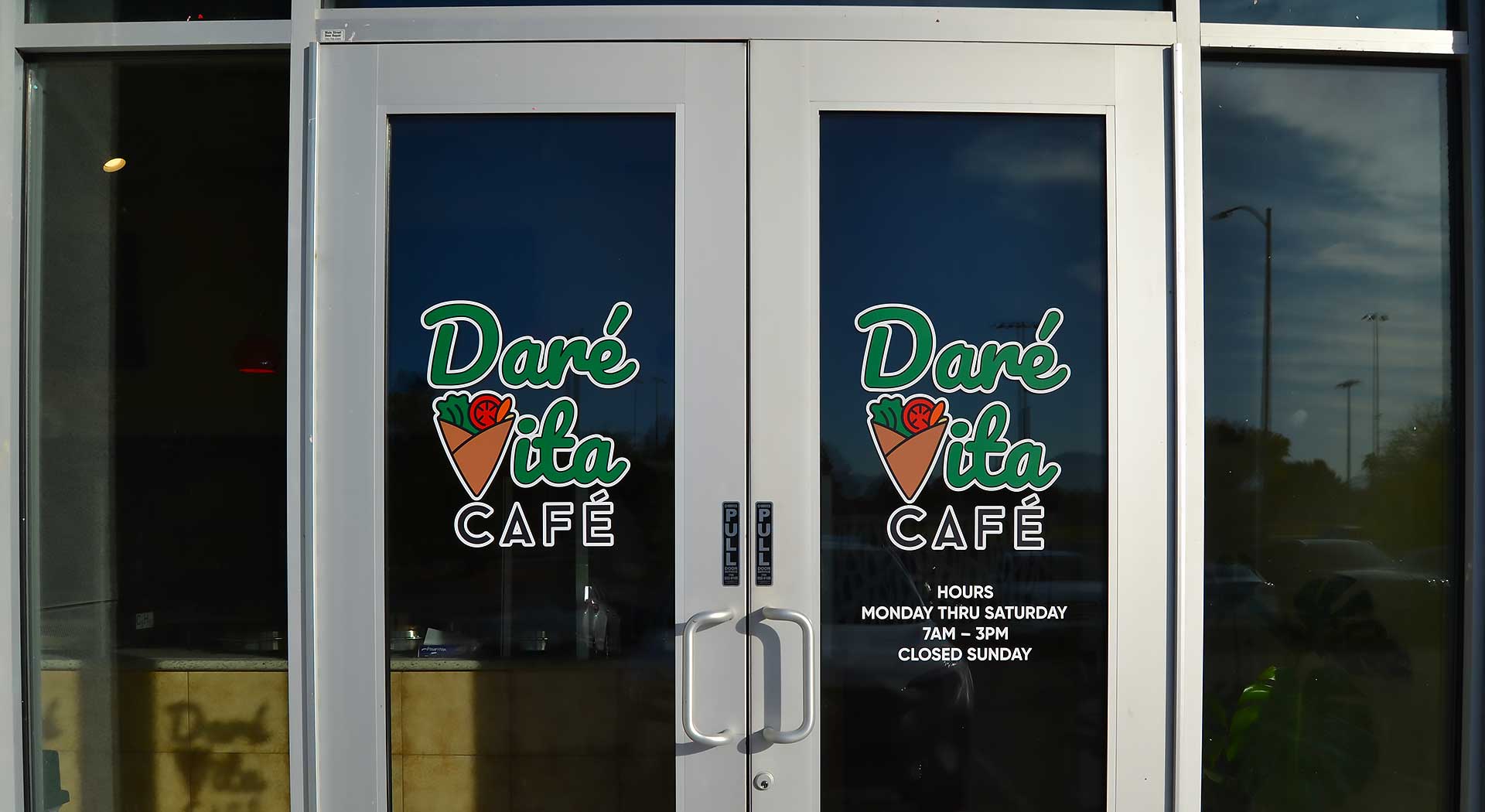 Entrance of Daré Vita Café with glass doors displaying the logo in green and white. The café offers allergen-friendly, plant-based meals, emphasizing fresh, wholesome ingredients. Visible hours indicate Monday to Saturday, 7 AM to 3 PM.| Daré Vita Café | Las Vegas