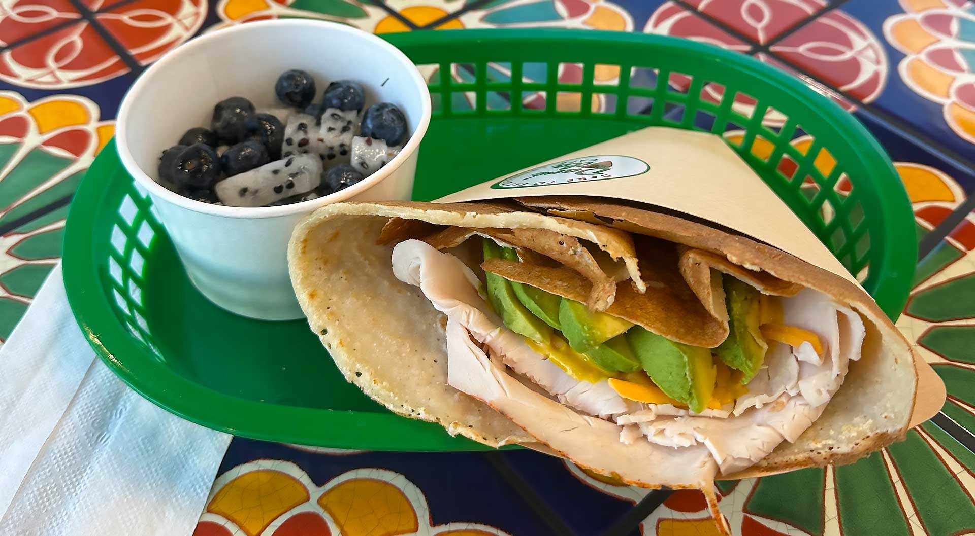 A vibrant, allergen-friendly crepe filled with turkey, avocado, and cheese sits in a green basket next to a white cup of fresh blueberries and dragon fruit, all on a colorful mosaic table, showcasing fresh, wholesome ingredients.| Daré Vita Café | Las Vegas